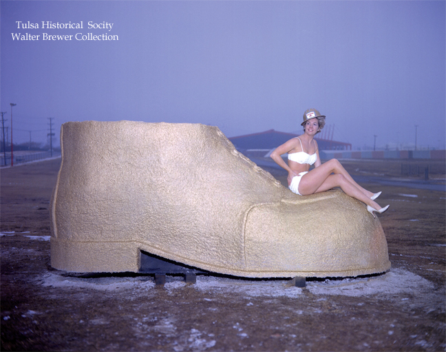 The golden driller Tulsa statue's shoe, circa 1950s. with swimsuit model sitting on it wearing a hardhat.