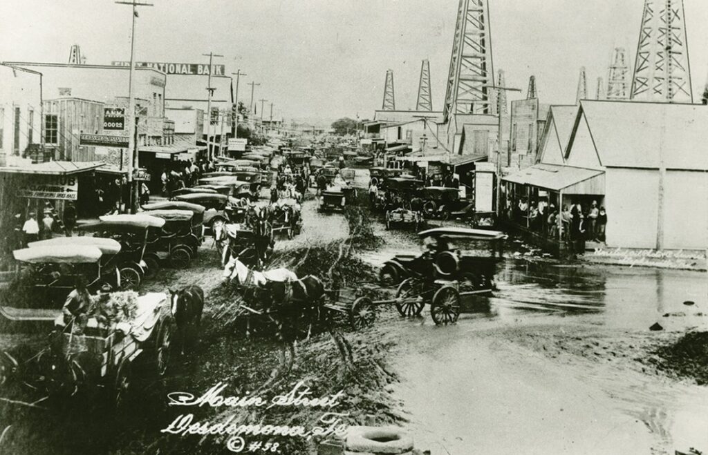 Autos in Desdemona, TX, stuck in muddy streets in front of oil derricks in 1918.